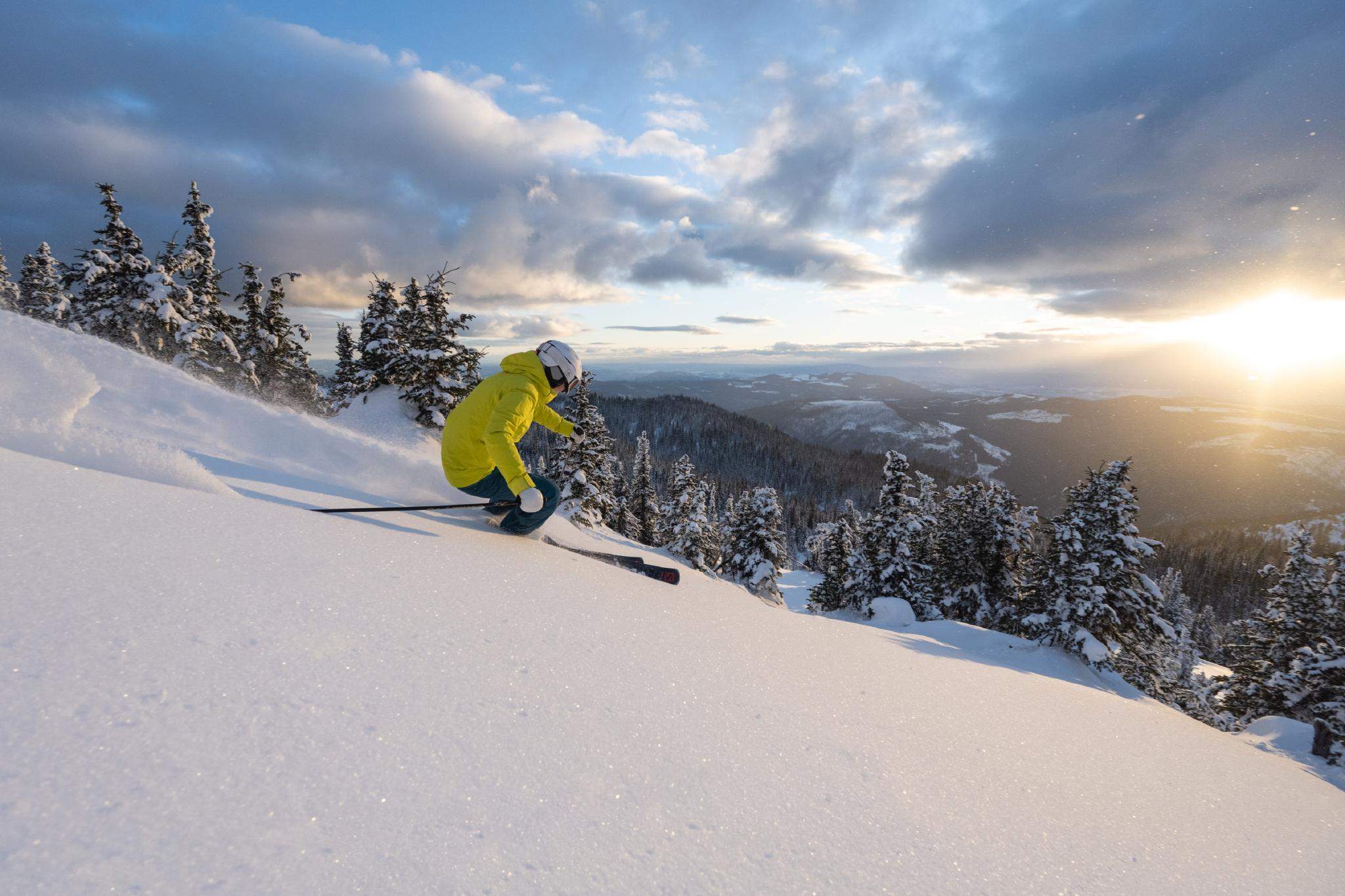 lone skier in a yellow jacket carving down a mountain in canada in powder with the sun setting over Sun Peaks