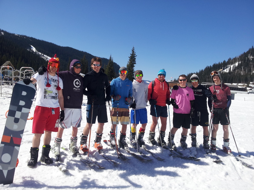 9 skiers in summer clothing with shorts behind snowy scene working abroad